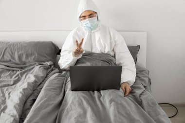 Koruyucu beyaz takım elbiseli ve tıbbi maskeli bir adam evde dizüstü bilgisayarlı bir yatakta çalışıyor ve web kamerasıyla arkadaşı veya iş arkadaşlarıyla konuşuyor. Coronavirüs salgını. Salgın sırasında uzak çalışma.