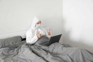 Koruyucu beyaz takım elbiseli ve tıbbi maskeli bir adam Coronavirus salgını yüzünden evde dizüstü bilgisayarla çalışıyor. Salgın sırasında uzaktan çalışma. COVID-19 karantina sırasında evde kal.