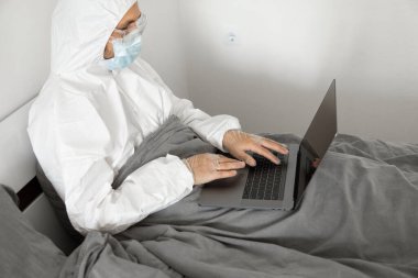 Koruyucu beyaz takım elbiseli ve tıbbi maskeli bir adam Coronavirus salgını yüzünden evde dizüstü bilgisayarla çalışıyor. Salgın sırasında uzaktan çalışma. COVID-19 karantina sırasında evde kal.