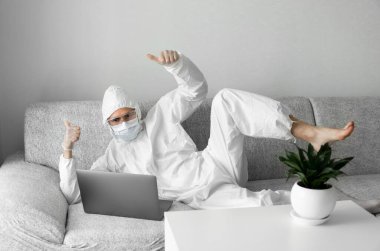 Koruyucu takım elbiseli ve tıbbi maskeli bir adam oturma odasındaki kanepede dizüstü bilgisayarla yatıyor ve projeden mutlu olduğu için iki parmağını kaldırıyor. Salgın COVID-19 karantinasında uzaktan çalışma.