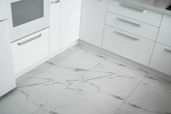 White lacquered kitchen facades on a comfortable kitchen with a marble kitchen tiles on the floor. Modern white kitchen clean interior design.