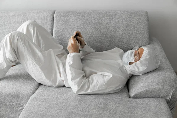 Homem de terno branco protetor e máscara médica está trabalhando em casa deitado em um sofá com um comprimido por causa da epidemia de coronavírus. Trabalho remoto durante a pandemia. Fique em casa durante a quarentena COVID-19 — Fotografia de Stock