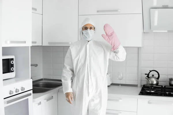 Man in white protective suit and in face medical mask and in a pink oven gloves cooks in the white kitchen at home during coronavirus. Stay at home. Enjoy cooking at home. COVID-19 global pandemic.