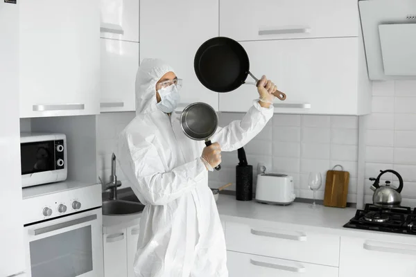 Man in white protective suit and in face medical mask holds a two frying pans in the white kitchen at home during coronavirus. Stay at home. Enjoy cooking at home. COVID-19 pandemic.