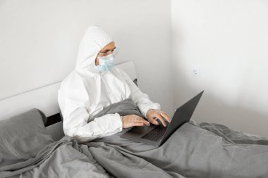 Koruyucu beyaz takım elbiseli ve tıbbi maskeli bir adam Coronavirus salgını yüzünden evde dizüstü bilgisayarla çalışıyor. Salgın sırasında uzaktan çalışma. COVID-19 karantina sırasında evde kal.