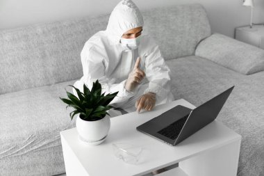 Koruyucu beyaz takım elbiseli ve tıbbi maskeli bir adam koronavirüs salgını nedeniyle evinde dizüstü bilgisayarla çalışıyor. Salgın sırasında uzaktan çalışma. COVID-19 karantina sırasında evde kal.