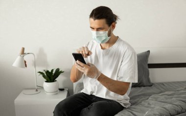 Maskeli bir adam evinde telefon kullanıyor. Coronavirüs salgını yüzünden yatağın üzerinde oturuyor. Salgın sırasında uzaktan çalışma. COVID-19 karantina sırasında evde kal.