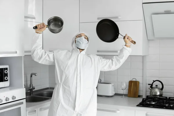 하얀 보호 복을 입고 얼굴에 의학용 마스크를 쓴 사람 이 코로나 바이러스가 유행하는 동안 집에 있는 흰 부엌에 프라이팬 두 개를 들고 있다. 집에 있어. 집에서 요리를 즐기 십시오. COVID-19 유행병. — 스톡 사진
