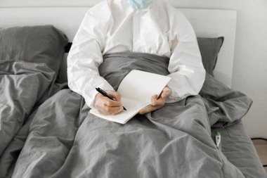 Korumacı beyaz takım elbiseli ve yüz maskeli bir adamın yatak odasında oturmasının nedeni 2019 yılının defterinde bir gün sayıp koronavirüs karantinasının sona ermesini beklemesi..