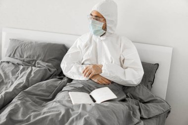 Korumacı beyaz takım elbiseli ve yüz maskeli bir adamın yatak odasında oturmasının nedeni 2019 yılının defterinde bir gün sayıp koronavirüs karantinasının sona ermesini beklemesi..