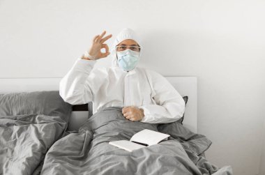 Korumacı beyaz takım elbiseli ve yüz maskeli bir adamın yatak odasında oturmasının nedeni 2019 yılının defterinde bir gün sayıp koronavirüs karantinasının sona ermesini beklemesi..