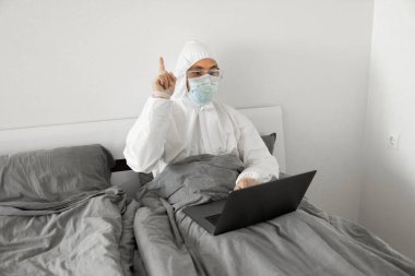 Koruyucu beyaz takım elbiseli ve tıbbi maskeli bir adam evde dizüstü bilgisayarlı bir yatakta çalışıyor ve web kamerasıyla arkadaşı veya iş arkadaşlarıyla konuşuyor. Coronavirüs salgını. Salgın sırasında uzak çalışma.