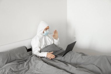 Koruyucu beyaz takım elbiseli ve tıbbi maskeli bir adam evde dizüstü bilgisayarlı bir yatakta çalışıyor ve Coronavirüs salgını nedeniyle web kamerasıyla arkadaşlarına veya iş arkadaşlarına merhaba diyor..