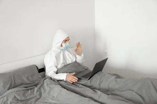 Férfi védő fehér öltöny és orvosi maszk dolgozik otthonról egy ágyban laptop és bemutatja helló barátai vagy üzleti partnerek webkamerával, mert a koronavírus járvány. — Stock Fotó