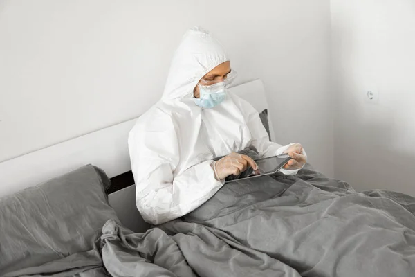 Man in protective white suit and medical mask is working from home in a bed with a tablet because of coronavirus epidemic. Remote work during pandemic. Stay home during COVID-19 quarantine concept.