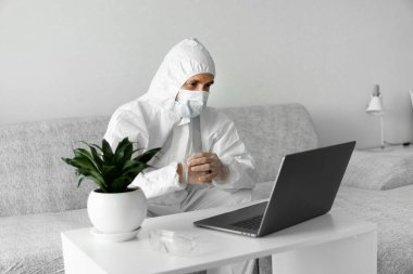 Koruyucu beyaz takım elbiseli ve tıbbi maskeli bir adam koronavirüs salgını nedeniyle evinde dizüstü bilgisayarla çalışıyor. Salgın sırasında uzaktan çalışma. COVID-19 karantina sırasında evde kal.