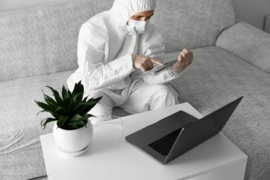 Koruyucu beyaz takım elbiseli ve tıbbi maskeli bir adam Coronavirus salgını nedeniyle evinde dizüstü bilgisayarı ve tableti olan bir koltukta çalışıyor. Salgın sırasında uzaktan çalışma. COVID-19 karantinası sırasında evde kal
