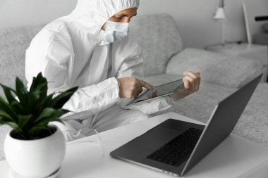Koruyucu beyaz takım elbiseli ve tıbbi maskeli bir adam Coronavirus salgını nedeniyle evinde dizüstü bilgisayarı ve tableti olan bir koltukta çalışıyor. Salgın sırasında uzaktan çalışma. COVID-19 karantinası sırasında evde kal
