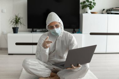 Koruyucu giysili ve tıbbi maskeli bir adam oturma odasında dizüstü bilgisayarla oturuyor çünkü Coronavirus yüzünden. Salgın sırasında uzaktan çalışma. COVID-19 karantinası sırasında evde kal. Serbest Çalışan.