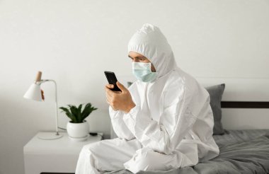 Koruyucu beyaz takım elbiseli ve tıbbi maskeli bir adam Coronavirüs salgını yüzünden evinde yatağında oturan bir telefonu kullanıyor. Salgın sırasında uzaktan çalışma. COVID-19 karantina sırasında evde kal.
