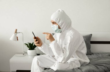Koruyucu beyaz takım elbiseli ve tıbbi maskeli bir adam Coronavirüs salgını yüzünden evinde yatağında oturan bir telefonu kullanıyor. Salgın sırasında uzaktan çalışma. COVID-19 karantina sırasında evde kal.