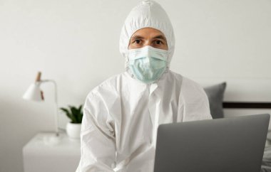 Koruyucu beyaz takım elbiseli ve tıbbi maskeli bir adam Coronavirus salgını yüzünden evde dizüstü bilgisayarla çalışıyor. Salgın sırasında uzaktan çalışma. COVID-19 karantina sırasında evde kal.