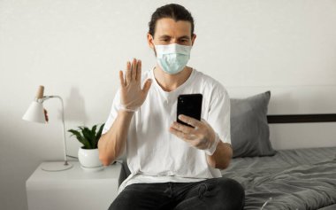 Tıbbi maskeli bir adam evinde bir telefon kullanıyor bir yatakta oturuyor ve web kamerasıyla arkadaşına ya da iş arkadaşına merhaba diyor. Coronavirüs salgını. Salgın sırasında uzak çalışma.