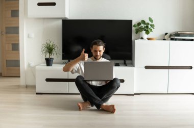 Sakallı bir adam, önünde dizüstü bilgisayarla çalışıyor ve web kamerasıyla arkadaşlarıyla, iş ortaklarıyla ya da müşterileriyle konuşuyor ve bir katta oturuyor. Evden çalışan serbest çalışan biri..