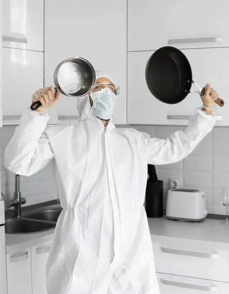 Man in white protective suit and in face medical mask holds a two frying pans in the white kitchen at home during coronavirus. Stay at home. Enjoy cooking at home. COVID-19 pandemic.