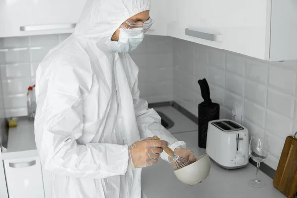 Man in white protective suit and in face medical mask cooks in the white kitchen at home during coronavirus. Stay at home. Enjoy cooking at home. COVID-19 global pandemic.