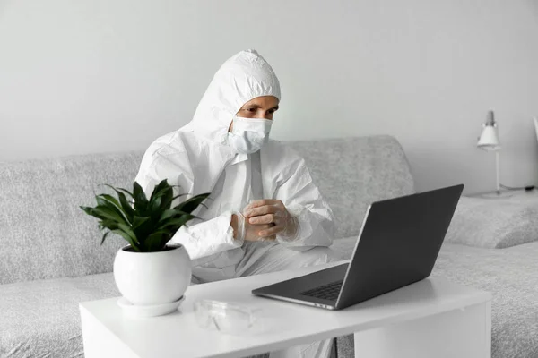 Man in protective white suit and medical mask is working from home on a sofa with laptop because of coronavirus epidemic. Remote work during pandemic. Stay home during COVID-19 quarantine concept.