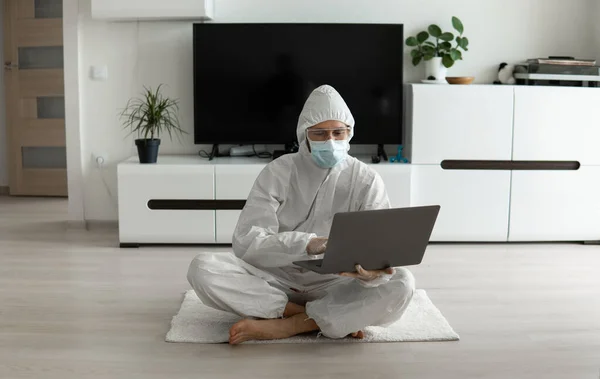 Man in protective suit and medical mask is sitting on a floor in his living room with a laptop because of coronavirus. Remote work during pandemic. Stay home during COVID-19 quarantine. Freelancer.