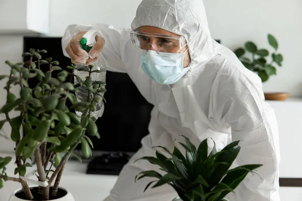 Man in protective suit, medical mask and rubber gloves is care about plants with a sprayer at his living room at home while coronavirus quarantine COVID-19.