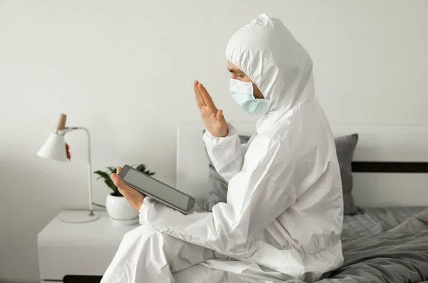 Man in protective white suit and medical mask is working from home on a bed with laptop and showing hello to his friends or business partners because of coronavirus epidemic. — Stock Photo, Image