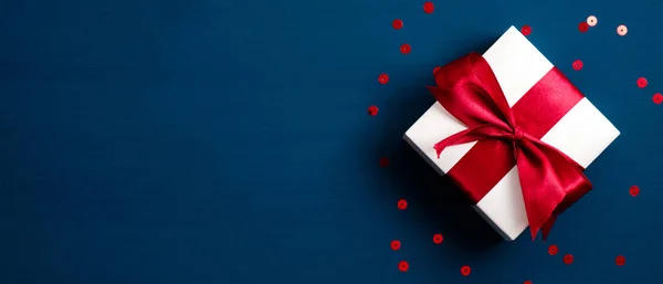 Białe pudełko z czerwoną wstążką na niebieskim tle z konfetti. Prezent świąteczny, niespodzianka walentynkowa, koncepcja urodzinowa. Płaskie ułożenie, widok z góry. — Zdjęcie stockowe