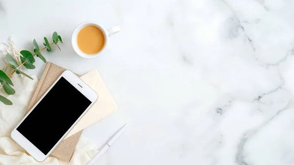 Akıllı telefon ekran modelli ofis masası, kağıt not defteri, bir fincan kahve, mermer arka planda okaliptüs yaprağı. Şık aksesuarları olan zarif kadın çalışma alanı. Düz yatış, üst görünüm.