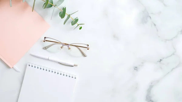 Vrouwelijke werkruimte met bril, papieren notitieblok, roze notitieboekje, pen, eucalyptusblad op marmeren tafel. Stijlvol thuiskantoor. — Stockfoto