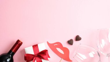 Şarap şişeli sevgililer günü masası, kırmızı kurdeleli beyaz hediye kutusu, gözlük, kalp şeklinde şekerler, pembe arka planda dudak süslemesi. En düşük düz konum biçimi kompozisyonu, üst görünüm