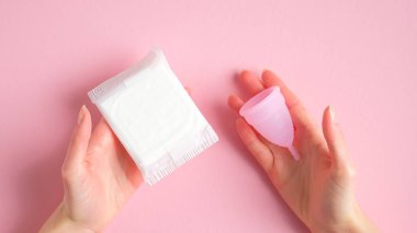 Hijyenik ped ve regl bardağını pembe arka planda tutan kadın elleri. Farklı türde regl hijyen ürünleri karşılaştırması. Kritik günler, adet döngüsü, kadın sağlık konsepti