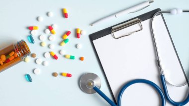 Doktor masasında tıbbi pano, steteskop, tablet ve haplar mavi arka planda cam bir şişeden dökülüyor. Düz yerde, üst tarafta. Eczane ve ilaç konsepti