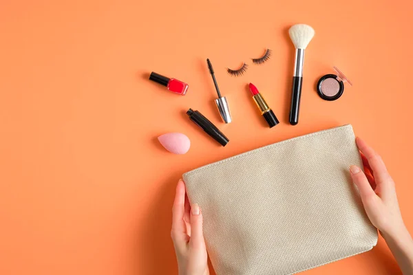 Female hands holding makeup bag with cosmetic products spilling out on to pastel peach color background. Flat lay, top view. Beauty salon banner template