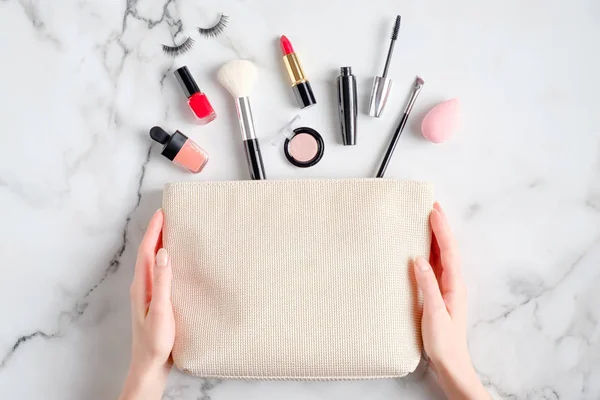 Female hands holding makeup bag with cosmetic products spilling out on to marble desk. Flat lay, top view
