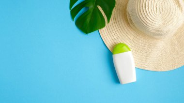 Güneş kremi şişesi, yeşil tropikal yaprak, mavi arka planda izole edilmiş kadın plaj şapkası. Güneş koruması, güneş kremi, cilt bakımı. Düz yatış, üst görünüm