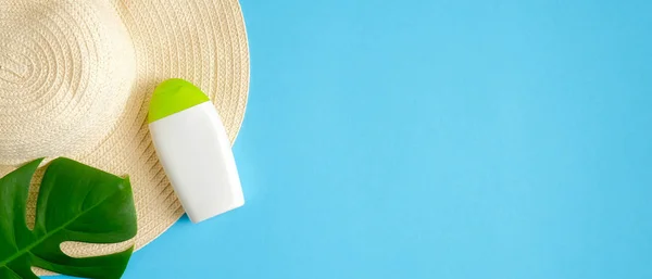 Sunscreen lotion bottle, female beach hat and tropical green leaf isolated on blue background. Sun protection and suntan cosmetic product concept. Flat lay, top view