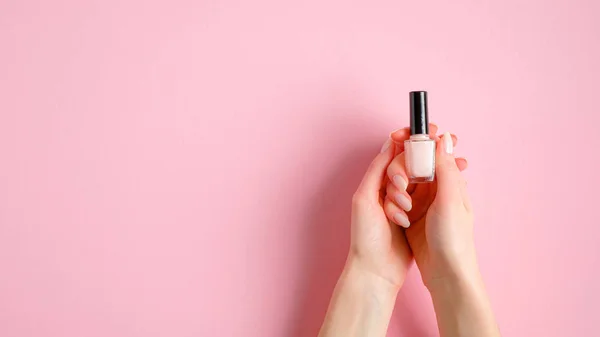 Nail polish bottle in female hands over pink background. Manicure, pedicure, beauty salon concept