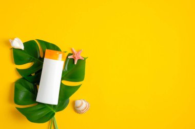 Etiketsiz boş güneş kremi modeli, sarı arka planda tropik yapraklar ve deniz kabukları. Güneş koruması ve cilt bakımı konsepti. Düz yatış, üst görünüm