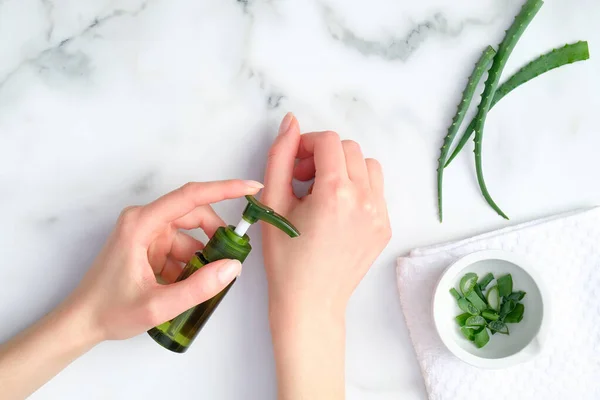 Cosmetic lotion bottle on female hands and green sliced stems aloe vera on white marble table. Woman applying organic moisturizing hand cream. Hand skin care concept