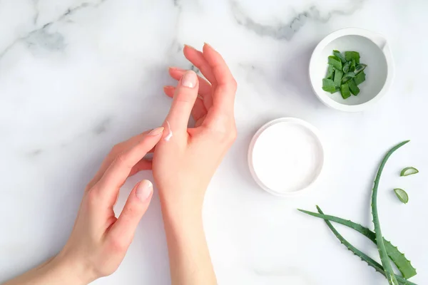 Kadın ellerinde kozmetik krem, mermer zemin üzerinde aloe vera kremi ve yeşil dilimlenmiş sapları olan kavanoz. Kadın organik nemlendirici el kremi sürüyor. El bakımı konsepti