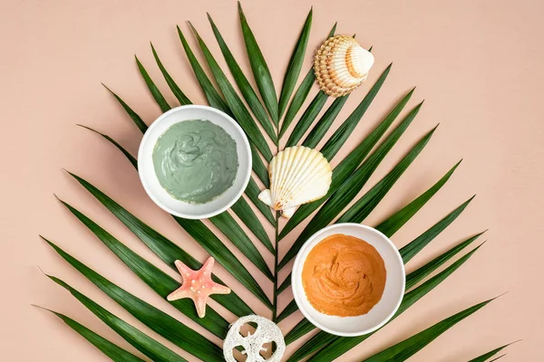 Cosmetic clay masks on peach color background with tropical palm leaf. SPA natural organic beauty products, summer facial skin care concept. Top view, flat lay.