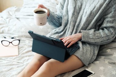 Tablet üzerinde çalışan ve evde yatakta kahve içen genç bir kadın. Samimi, kızlık zarı tarzı. Çevrimiçi alışveriş, çalışma evi, serbest çalışma, çevrimiçi öğrenme, öğrenim konsepti. Uzaklık eğitimi